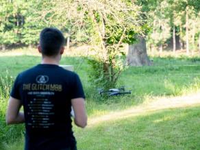 Blick von hinten auf einen Mann, der eine Drohne steuert, die etwa einen Meter über einer Wiese fliegt. Im Hintergrund Wald.