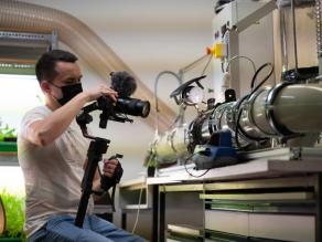 Ein Kameramann filmt einen Glas-Windkanal mit etwa 25 cm Durchmesser, an den diverse Kabel und Instrumente angeschlossen sind.