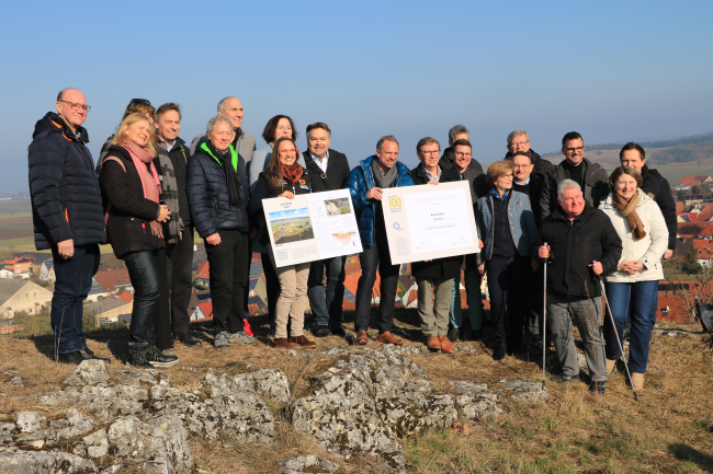 Eine Gruppe von 20 Personen steht auf einer felsigen Kuppe. In der Bildmitte wird die stark vergrößerte Kopie einer Urkunde gezeigt.