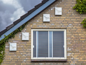 An der ockerfarbenen Klinkerfassade eines Wohnhauses hängen Fledermauskästen über und neben einem Fenster. Links und rechts rankt eine Kletterplanze die Wand empor bis zum Dach.