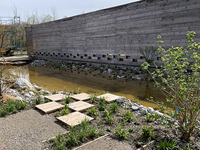Blick auf die Vorderseite des Pavillons und den davor liegenden Teich. Die Wand besteht aus geschichteten Holzbalken.
