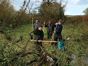 Fünf Jugendliche stehen mit Wathosen in und an einem Bach und legen mit Harken das Bachbett zur Gewährleistung des Abflusses frei. Die Ufer sind dicht mit Gras und Schilf bewachsen. Eine Totholzstruktur befindet sich im Vordergrund am Bachufer.