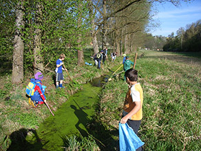 Mehrere Kinder stehen mit Harken und Müllsäcken am Ufer eines Bachs.