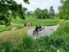 Zwei Personen sitzen auf einer Bank mit Blick auf eine Niederung. Im Hintergrund ist ein Bach zu erkennen. Eine Menschengruppe steht am Ufer.