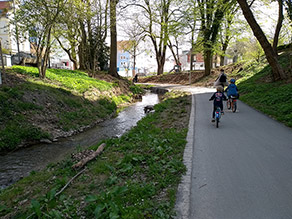 Eine Frau und zwei Kinder fahren mit dem Fahrrad auf einem Geh- und Radweg entlang eines Bachs.