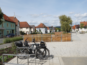 Fahrradständer mit zwei Elektro-Fahrrädern vor einem Geländer mit Holzverkleidung. Im Hintergrund ist das neue Gewässerbett zu erahnen.