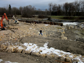 Ein Bauarbeiter und zwei Bagger bei Steinsatzarbeiten im Bereich der neuen Sohlrampe. Im Hintergrund ist der durch Spundwände vom Baufeld abgetrennte Fluss zu erkennen.