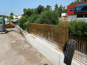 Eine Hochwasserschutzmauer am Ufer eines Flusses befindet sich im Bau. Stahlmatten stehen aus einer niedrigen Betonmauer heraus. Im Hintergrund lagerndes Baumaterial.