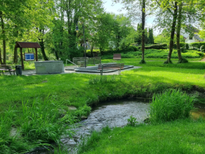 Ein Bach fließt durch eine mit Bäumen bestandenen Wiese in einer Grünanlage. Im Hintergrund eine Kneippanlage mit Kneippbecken, Armtauchbecken, Holzbank und Infotafel.