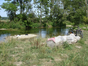 Am flachen Ufer eines Flusses liegt ein großer Baumstamm mit einem Handtuch darauf. Daneben stehen zwei Fahrräder. Auf einer kleinen Kiesinsel im Wasser liegen ein Rucksack, Handtuch und Schuhe.