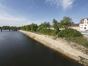 Das Ufer eines Flusses wird neugestaltet. Im Bereich der Böschung entsteht ein Geh- und Radweg mit niedriger Natursteinmauer.