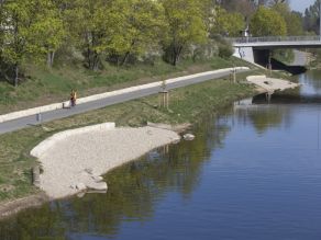 Eine halbmondförmige Kiesfläche mit Sitzsteinreihe am Ufer eines Flusses. Auf dem dahinter parallel zum Fluss verlaufenden Geh- und Radweg fährt eine Person Fahrrad.