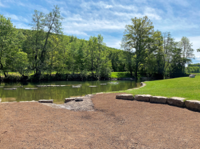 Ein neu hergestelltes Becken wird durch Steinquader von den anschließenden Grasflächen begrenzt und öffnet sich hin zu einem Fluss mit einem Flachwasserbereich und Trittsteinen im Wasser.