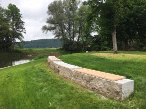 Auf einer grasbewachsenen Uferböschung eines Flusses eine Sitzmauer aus Steinquadern mit Holzauflage.