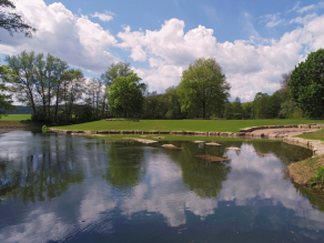 Ein Fluss mit Flachwasserbereich, dessen Ufer mit Steinquadern befestigt sind und zum Sitzen am Wasser einladen. Dahinter schließen sich Wiesenflächen mit Sitzmauern an.