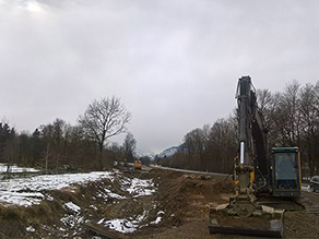 Das Gelände im Bereich des Hochwasserschutz Freilassing während der Baumaßnahme: Zwischen einer Straße und dem Mühlbach im neuen Flussbett eine schneebedeckte Fläche und das alte Flussbett. Rechts davon ein Bagger.