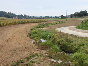 Kleiner Bach mit erstem Uferbewuchs nach der Maßnahmenumsetzung. Links davon schließt sich eine Fläche ohne Bewuchs und ein Maisfeld an. Rechts des Baches verläuft ein neuer Geh- und Radweg.