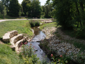 Eine Sitzstufenanlage aus Natursteinen in der Böschung eines Bachs, der leicht geschwungen durch eine Grünanlage fließt. Parallel zum Gewässer verläuft ein Geh- und Radweg.