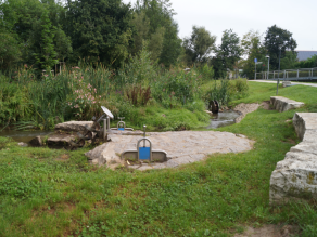 Ein Wasserspielbereich für Kinder mit Archimedischer Schraube, Stauwehren und Wasserrad an einem kleinen Bach.  Daneben bieten zwei Steinmäuerchen Sitzmöglichkeiten am Ufer.