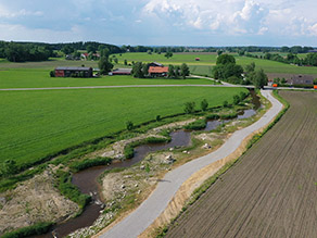 Ein Geh- und Radweg verläuft parallel zu einem renaturierten Bach mit geschwungenem Verlauf durch eine landwirtschaftlich geprägte Landschaft.