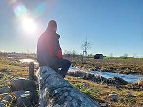 Ein Mann sitzt auf einem entrindeten Baumstamm am Ufer eines Baches. Die Sonne spiegelt sich im Wasser des leicht geschwungen fließenden Baches. Im Hintergrund landwirtschaftliche Flächen und eine Hütte.