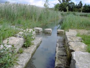 Die Ufer eines kleinen Bachs sind mit Natursteinquadern befestigt und bieten Sitzmöglichkeiten am Wasser. Im Hintergrund führen Trittsteine über den Bach.