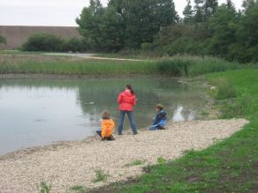Drei Kinder stehen und hocken am flachen Kiesufer eines Gewässers und werfen Steine in das Wasser.