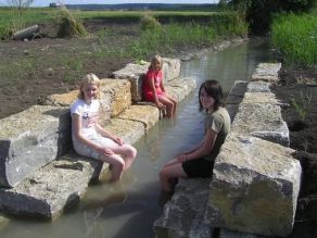 Drei Kinder sitzen auf Natursteinquadern am Ufer eines Bachs mit ihren Beinen im Wasser.