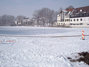 Eine ausgedehnte Fläche mit Schneedecke vor Wohngebäuden im Hintergrund.