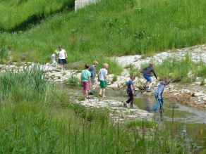 Mehrere Kinder spielen an einem Bach mit abgeflachten, mit Steinsatz gesicherten Ufern. Es schließt sich eine grasbewachsene Böschung an.