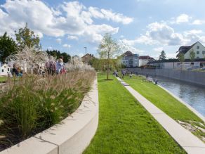 Eine Grünanlage an einem Fluss im städtischen Raum mit Wiesenflächen und mehreren Sitzreihen.