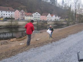 Zwei Personen verteilen Saatgut auf einer Uferböschung am Fluss.