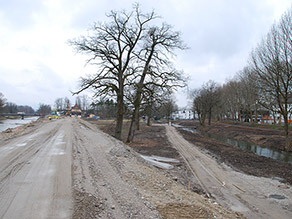 Blick vom Hochwasserschutzdeich während der Umbaumaßnahmen auf den Bereich zwischen Mangfall und Hammerbach (Mangfallpark Süd). Eine Baustraße führt entlang des Hammerbachs. Im Hintergrund stehen Baumaschinen und Kraftfahrzeuge auf dem brach liegenden Deich.
