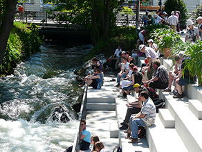 Am Zusammenfluss des Mühlbachs und des Hammerbachs am Ichikawaplatz strömt Wasser über Felsen und zahlreiche Personen sitzen daneben durch ein Geländer getrennt auf Sitzstufen in der Sonne.