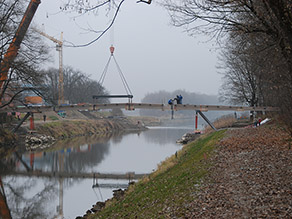 Über einen Fluss wird das Element einer neuen Fußgängerbrücke eingehoben. Mehrere Personen sind mit der Montage auf der Brücke beschäftigt.