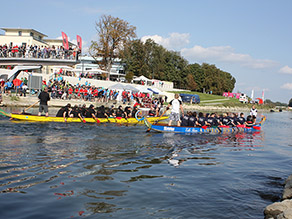 Auf einem Fluss zwei bunte Drachenboote mit mehreren Personen darin. Im Hintergrund eine große Menschenmenge am Flussufer und auf einem Aussichtsteg.