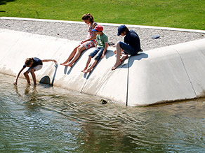 Auf einem zum Wasser abfallenden Gestaltungselement aus Beton an einem Bachufer sitzen mehrere Personen. Ein Kind steigt in das flache Wasser im Randbereich.