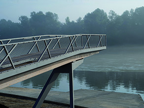 Ein Aussichtssteg mit Geländer ragt über das betonierte Ufer eines Flusses, dessen gegenüberliegendes Ufer im Hintergrund im Nebel verschwindet.