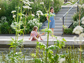 Blick durch Uferbepflanzung auf das gegenüberliegende Ufer eines Kanals. Auf der Ufermauer sitzt ein Kind, dahinter kommt eine Person eine Treppe herunter. Die Uferböschungen sind mit Blumenbeeten bepflanzt.