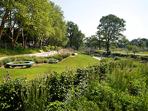 Am Ufer eines Gewässers eine Wiesenfläche und Blumenbeete mit diversen Gestaltungselementen. Gehwege führen parallel zum Gewässer durch die Grünanlage.