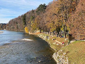 Blick von der Isarbrücke in Bad Tölz flussabwärts auf das Taubenloch unter herbstlichen Bedingungen. Mehrere Personen sitzen auf Mauern, die in die Uferböschung eingebunden sind, sowie auf Holzbänken.