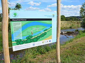 Eine Infotafel mit Holzpfosten steht am Ufer eines Flusses und informiert über die Wasserinsel Alzenau.
