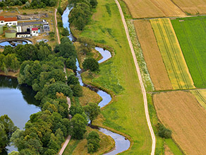Luftaufnahme der renaturierten Kahl flussaufwärts, die sich angrenzend an Wiesenflächen und von Geh- und Radwegen begleitet durch die Landschaft schlängelt. Neben den Wegen Seen und Ackerflächen.