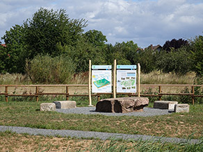 Zwei Infotafeln zum Wasserkreislauf und zu der Kahl stehen vor einem Holzgeländer. Davor einige Sitzmöglichkeiten aus Stein und ein Kiesweg.