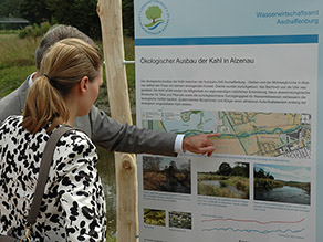 Zwei Personen vor einer Infotafel zum Ökologischen Ausbau der Kahl in Alzenau, die am Ufer eines Flusses steht.
