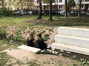 Auf einer Treppe sitzen zwei Mädchen am Ufer des Bachs. Sitzstufen am gegenüberliegenden Ufer ermöglichen das Verweilen am Wasser.