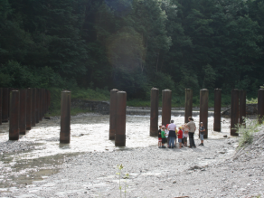 Bei einem Aktionstag zum Wildbachlehrpfad steht eine Gruppe von Kindern mit mehreren Erwachsenen am Ufer eines Wildbachs vor einem Wildholzrechen aus Stahlrohren. Im Hintergrund strömt Wasser über eine Wildbachsperre.