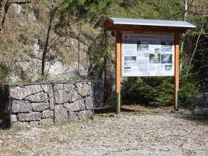Eine Infotafel des Wildbachlehrpfades am Lainbach zu den Bauwerken der Wildbachverbauung (Lehrtafel-Nr. 8) steht vor einem Abhang. Daneben ein anschauliches Exemplar eines Drahtschotterkastens.