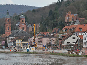 Blick über einen Fluss hinweg auf eine Baustelle am gegenüberliegenden Ufer. Im Hintergrund die Altstadt Miltenbergs mit Wohngebäuden und Kirchtürmen.