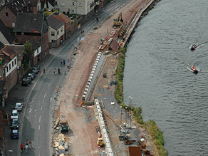 Luftaufnahme einer Baustelle zwischen einem Fluss und einer Straße mit angrenzender Wohnbebauung während der Herstellung von Hochwasserschutzmauern.